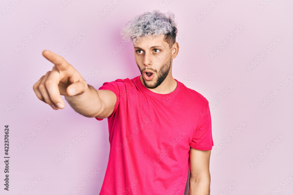 Young hispanic man with modern dyed hair wearing casual pink t shirt pointing with finger surprised ahead, open mouth amazed expression, something on the front Young hispanic man with modern dyed hair wearing casual pink t shirt pointing with finger surprised ahead, open mouth amazed expression, something on the front