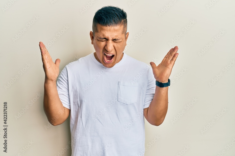 Fototapeta premium Young latin man wearing casual white t shirt celebrating mad and crazy for success with arms raised and closed eyes screaming excited. winner concept