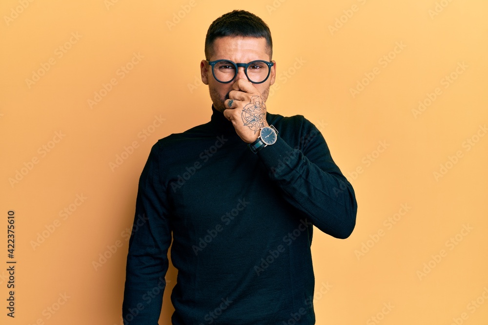 Handsome man with tattoos wearing turtleneck sweater and glasses smelling something stinky and disgusting, intolerable smell, holding breath with fingers on nose. bad smell Handsome man with tattoos wearing turtleneck sweater and glasses smelling something stinky and disgusting, intolerable smell, holding breath with fingers on nose. bad smell