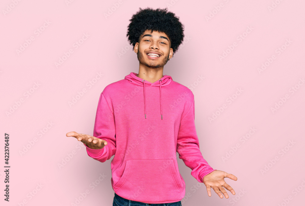 Young african american man with afro hair wearing casual pink sweatshirt smiling cheerful with open arms as friendly welcome, positive and confident greetings Young african american man with afro hair wearing casual pink sweatshirt smiling cheerful with open arms as friendly welcome, positive and confident greetings