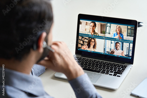 Indian business man wearing headset having virtual team meeting on video conference call using laptop work from home office talking to diverse group in remote online distance chat. Over shoulder view