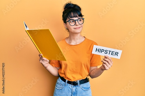 Young hispanic girl reading book and holding paper with hipster message smiling looking to the side and staring away thinking.