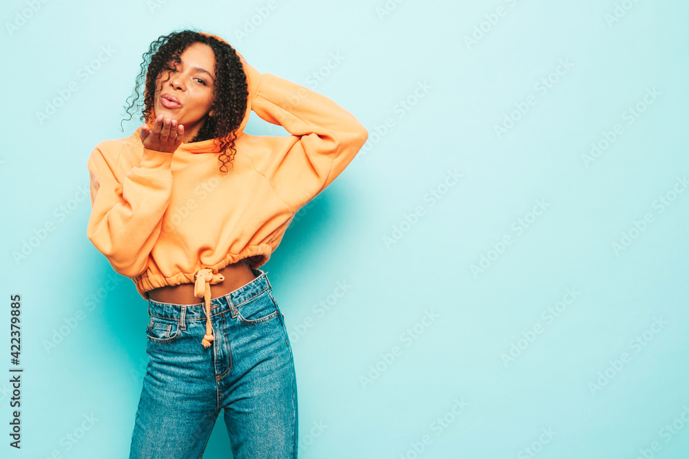 Beautiful black woman with afro curls hairstyle.Smiling model in orange hoodie and trendy jeans clothes. Sexy carefree female posing near blue wall in studio in sunglasses. Gives air kiss Beautiful black woman with afro curls hairstyle.Smiling model in orange hoodie and trendy jeans clothes. Sexy carefree female posing near blue wall in studio in sunglasses. Gives air kiss