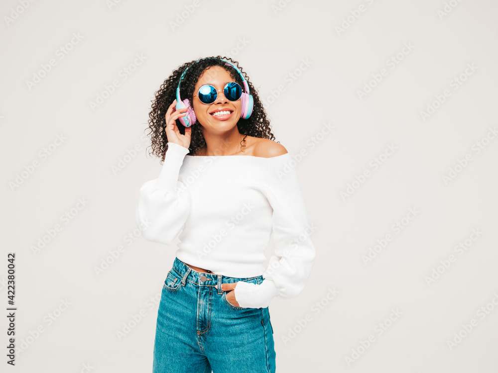 Beautiful black woman with afro curls hairstyle.Smiling model in sweater and jeans.Sexy carefree female listening music in wireless headphones.Posing in studio on white background in sunglasses Beautiful black woman with afro curls hairstyle.Smiling model in sweater and jeans.Sexy carefree female listening music in wireless headphones.Posing in studio on white background in sunglasses