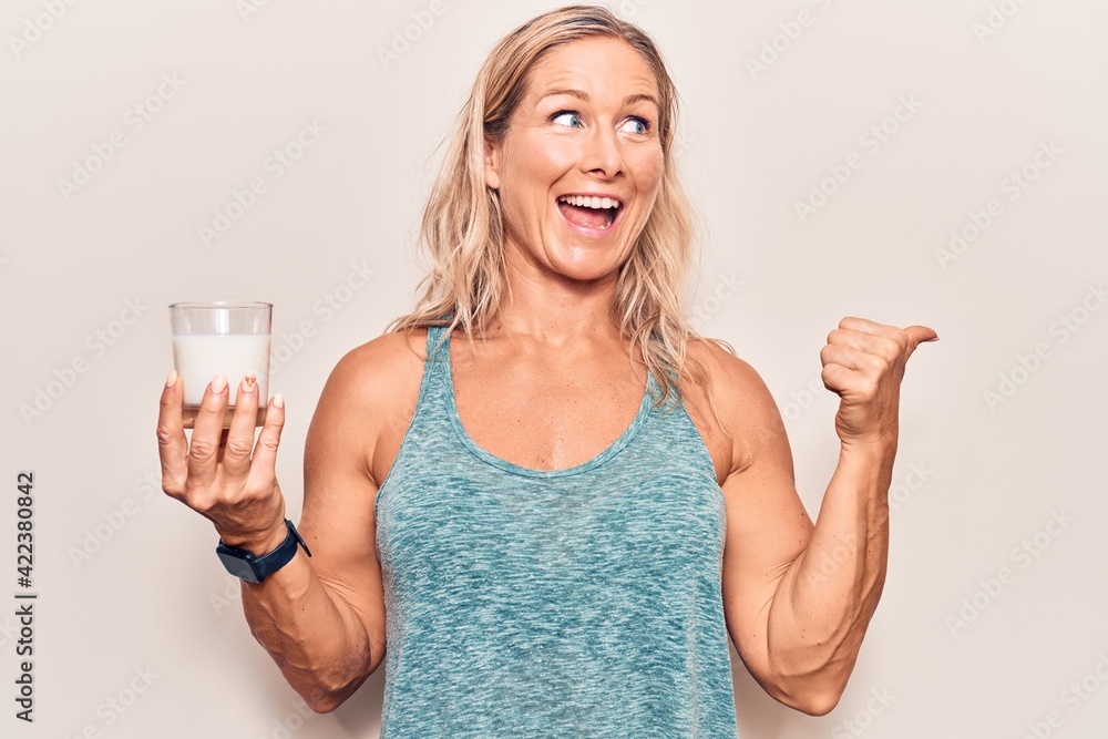 Middle age caucasian blonde woman holding a glass of milk pointing thumb up to the side smiling happy with open mouth