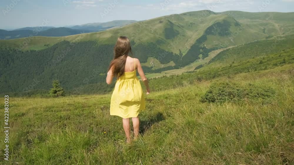 Rear view of a happy woman hiker in yellow dress standing on grassy hill on a windy day in summer mountains with outstretched arms enjoying view of nature. Rear view of a happy woman hiker in yellow dress standing on grassy hill on a windy day in summer mountains with outstretched arms enjoying view of nature.