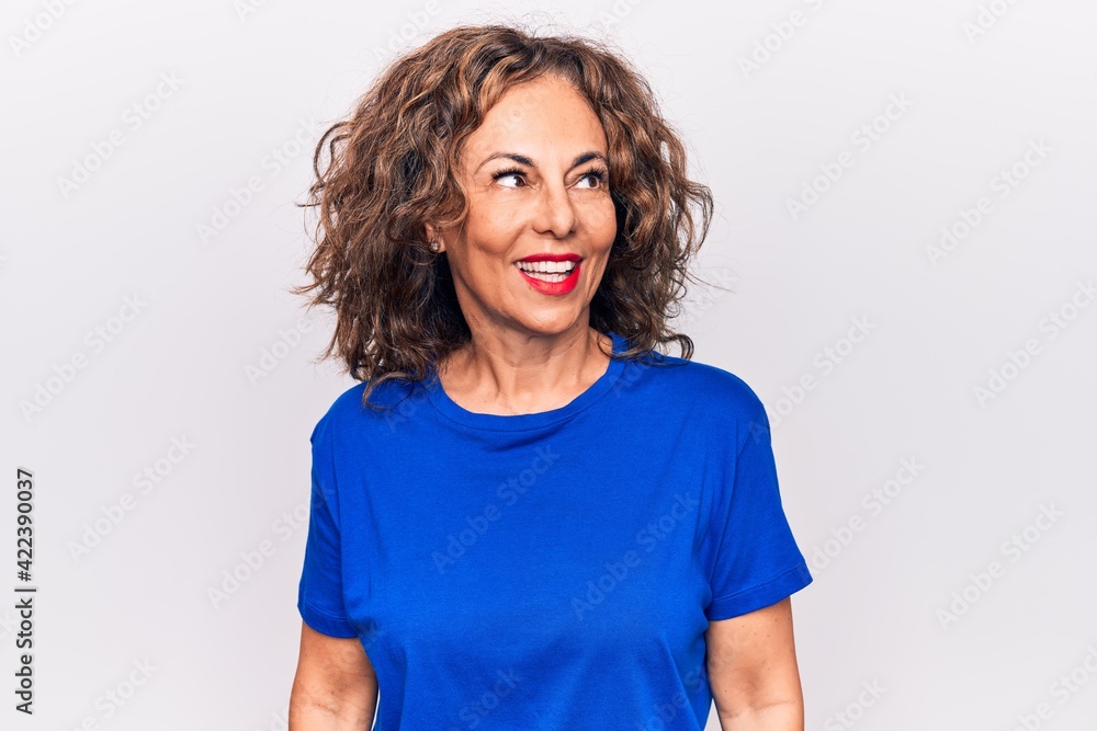 Middle age beautiful brunette woman wearing blue t-shirt standing over white background looking to side, relax profile pose with natural face and confident smile. Middle age beautiful brunette woman wearing blue t-shirt standing over white background looking to side, relax profile pose with natural face and confident smile.