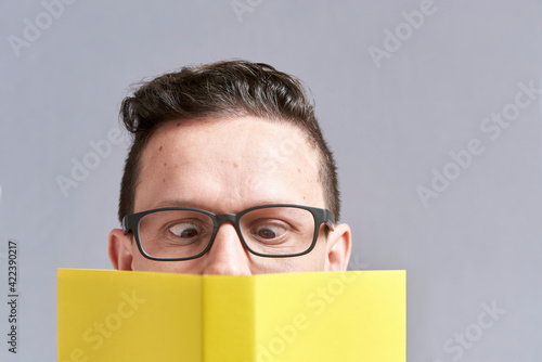 Young man wearing glasses reading a yellow book with squinted eyes; a funny look