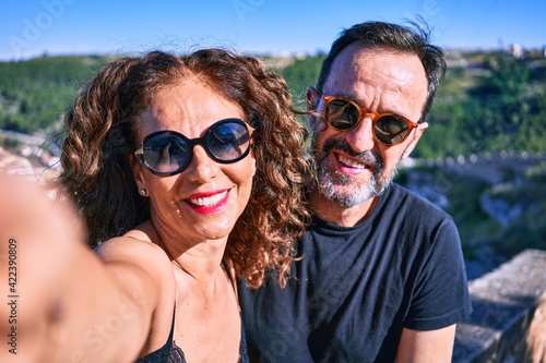 Middle age beautiful couple wearing casual clothes and sunglasses. Sitting on stone wall making selfie by the camera over town landscape background