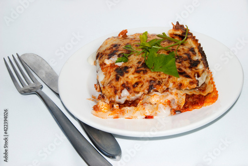 Portion of lasagna bolognese on white plate and covered with knife and fork on white background