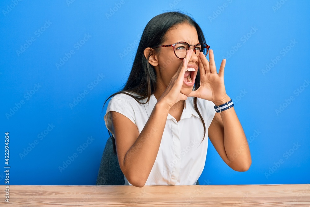 Beautiful hispanic woman wearing casual clothes sitting on the table shouting angry out loud with hands over mouth Beautiful hispanic woman wearing casual clothes sitting on the table shouting angry out loud with hands over mouth