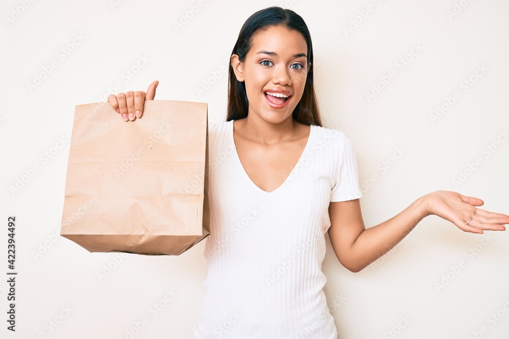 Young beautiful latin girl holding take away paper bag celebrating achievement with happy smile and winner expression with raised hand Young beautiful latin girl holding take away paper bag celebrating achievement with happy smile and winner expression with raised hand