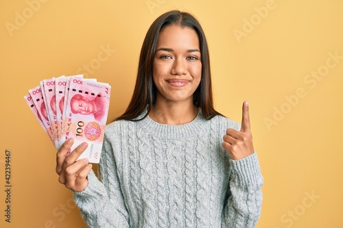 Beautiful hispanic woman holding 100 yuan chinese banknotes smiling with an idea or question pointing finger with happy face, number one