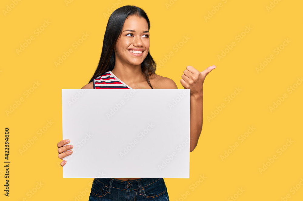 Beautiful hispanic woman holding blank empty banner pointing thumb up to the side smiling happy with open mouth