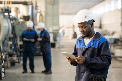Young African male factory worker in overalls and hardhat using digital tablet while standing in front of camera against two busy colleagues
