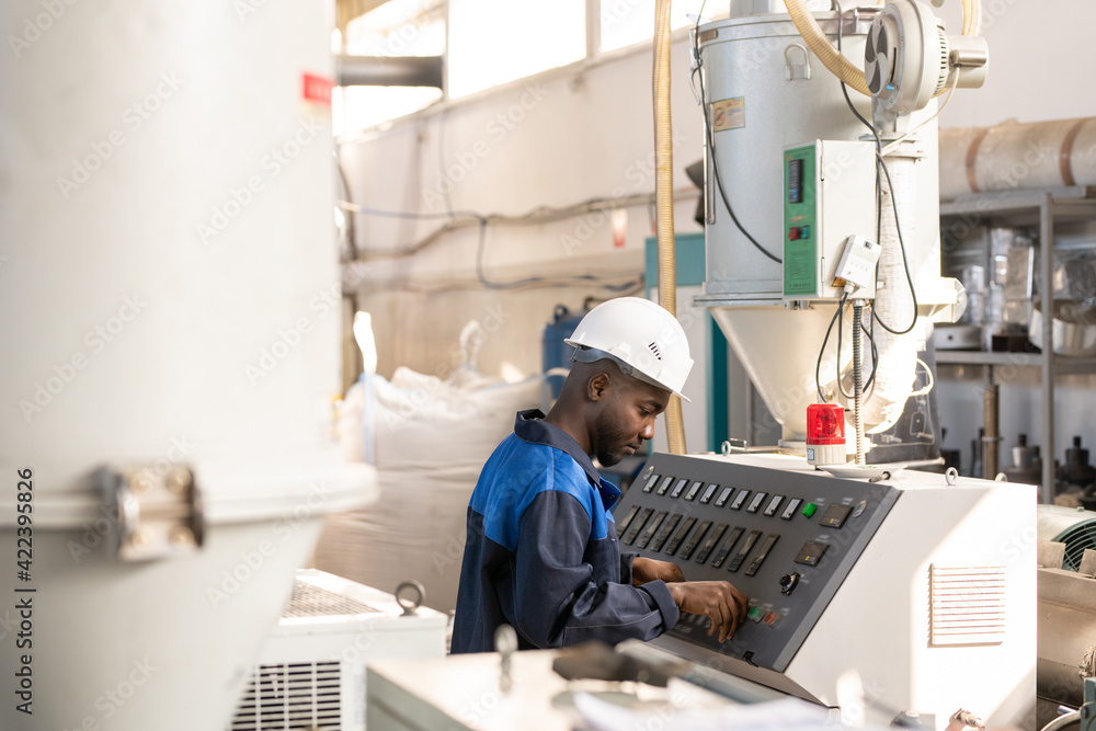 Side view of young African worker in overalls and hardhat standing in front of control panel of huge industrial machine and adjusting mechanism Side view of young African worker in overalls and hardhat standing in front of control panel of huge industrial machine and adjusting mechanism