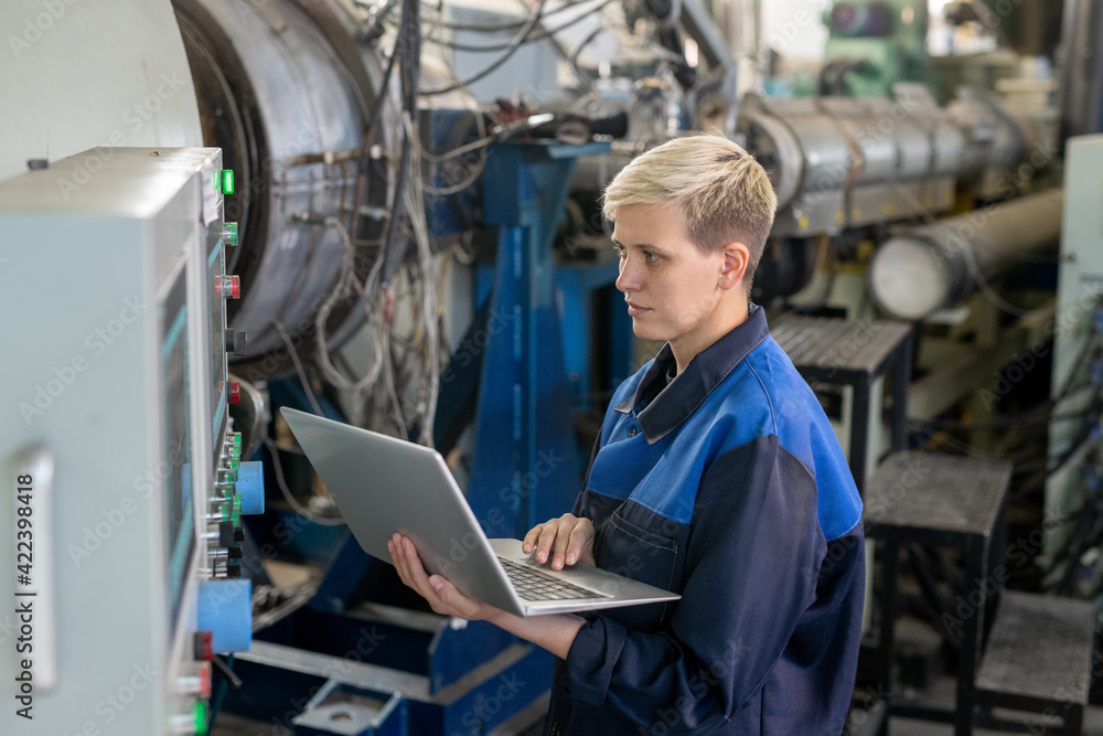 Young serious blond female worker in overalls using laptop while looking through online technical data against industrial equipment in workshop