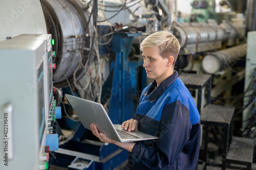Young serious blond female worker in overalls using laptop while looking through online technical data against industrial equipment in workshop © pressmaster