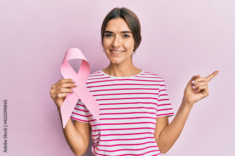 Young hispanic woman holding pink cancer ribbon smiling happy pointing with hand and finger to the side Young hispanic woman holding pink cancer ribbon smiling happy pointing with hand and finger to the side