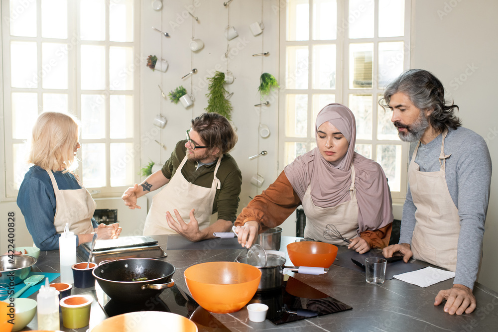 Young Muslim female trainee in hijab holding whisker over pan with water on electric stove while cooking at masterclass among other learners Young Muslim female trainee in hijab holding whisker over pan with water on electric stove while cooking at masterclass among other learners
