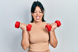 Young hispanic woman wearing sportswear using dumbbells sticking tongue out happy with funny expression.