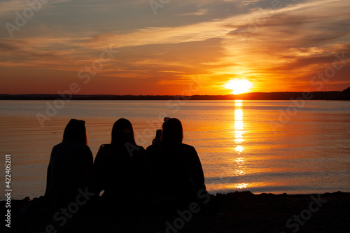 Horizontal landscape photography with a three sitting and chilling on the beach friend silhouettes against colorful sky with setting sun during calm sunset 