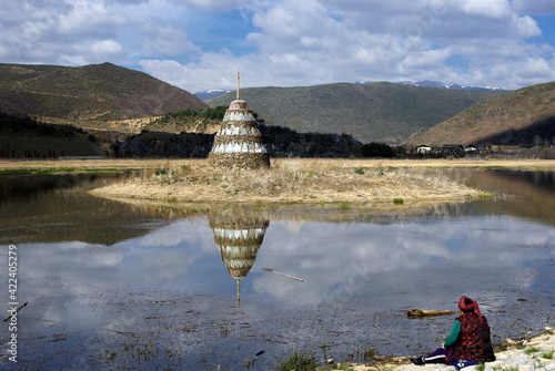 A Tibetan woman sits on the shore of Lamuyangcuo Lake with a Buddhist stupa on a small island, Shangri-la, Yunnan Province, China. photo