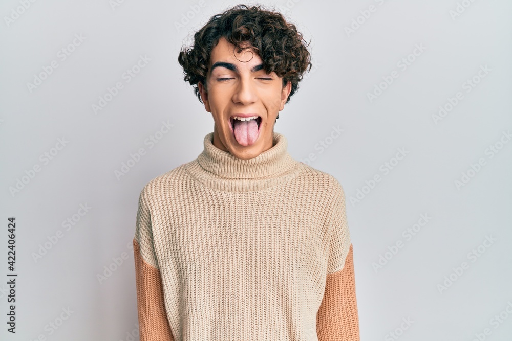 Hispanic young man wearing casual winter sweater sticking tongue out happy with funny expression. emotion concept. Hispanic young man wearing casual winter sweater sticking tongue out happy with funny expression. emotion concept.