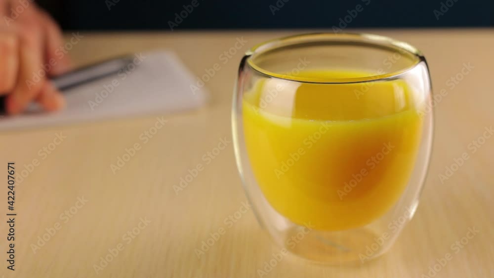 Glass with orange juice in the foreground. The woman writes tasks in a notebook in the background and periodically picks up a cup of drink with his hand. Glass with orange juice in the foreground. The woman writes tasks in a notebook in the background and periodically picks up a cup of drink with his hand.