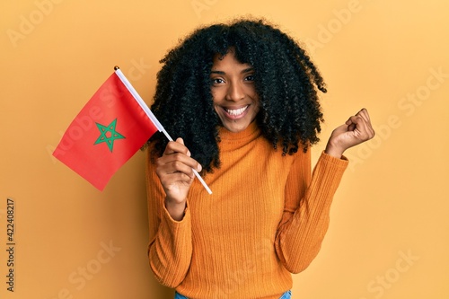 African american woman with afro hair holding morocco flag screaming proud, celebrating victory and success very excited with raised arm