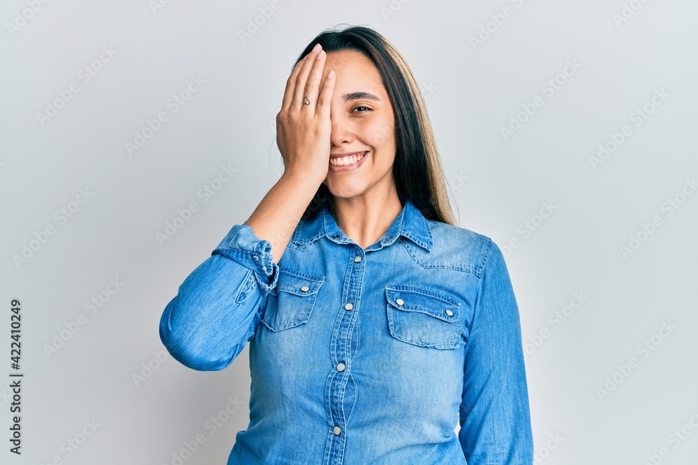 Young hispanic woman wearing casual denim jacket covering one eye with hand, confident smile on face and surprise emotion. Young hispanic woman wearing casual denim jacket covering one eye with hand, confident smile on face and surprise emotion.
