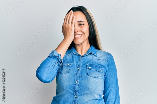 Young hispanic woman wearing casual denim jacket covering one eye with hand, confident smile on face and surprise emotion.