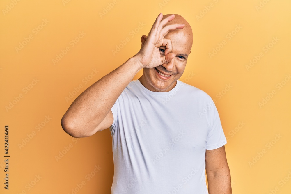 Middle age bald man wearing casual white tshirt smiling happy doing ok sign with hand on eye looking through fingers Middle age bald man wearing casual white tshirt smiling happy doing ok sign with hand on eye looking through fingers