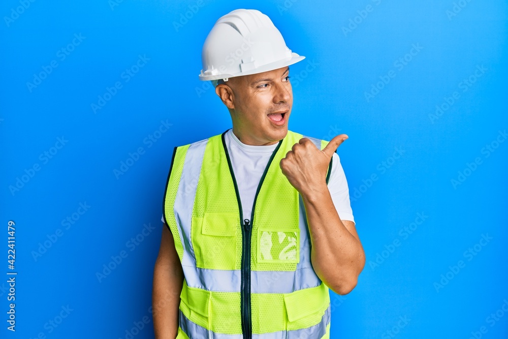 Middle age bald man wearing architect hardhat pointing thumb up to the side smiling happy with open mouth