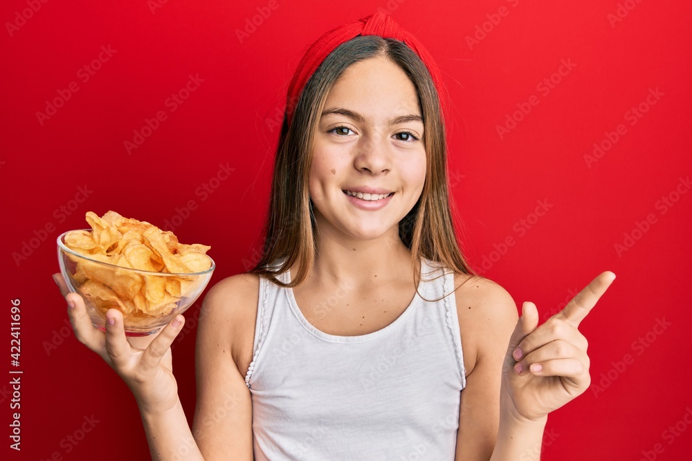 Beautiful brunette little girl holding potato chips smiling happy pointing with hand and finger to the side Beautiful brunette little girl holding potato chips smiling happy pointing with hand and finger to the side