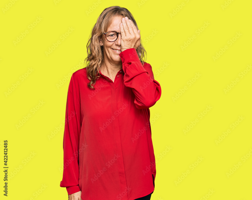 Middle age blonde woman wearing casual shirt over red background covering one eye with hand, confident smile on face and surprise emotion. Middle age blonde woman wearing casual shirt over red background covering one eye with hand, confident smile on face and surprise emotion.