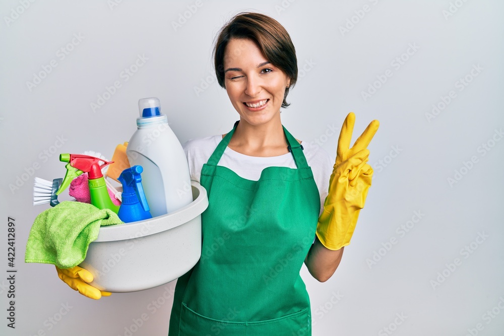 Young brunette woman with short hair wearing apron holding cleaning products smiling with happy face winking at the camera doing victory sign with fingers. number two. Young brunette woman with short hair wearing apron holding cleaning products smiling with happy face winking at the camera doing victory sign with fingers. number two.