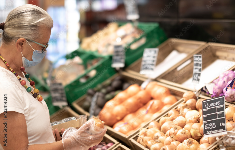A senior retired woman white dressed  wearing surgical mask due to coronavirus shopping and choosing vegetables in a store using protective gloves