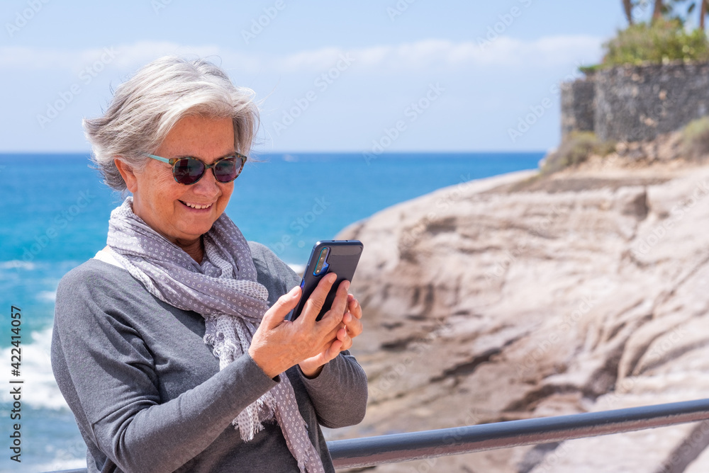 Attractive senior woman in sea vacation, using phone and smiling. Blue sea and horizon over water