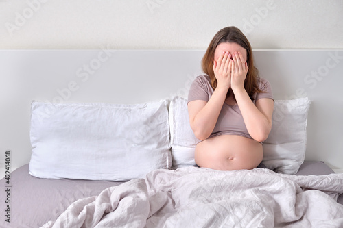 A lonely pregnant woman lies in bed covering face with her hands. Pregnancy and loneliness problems