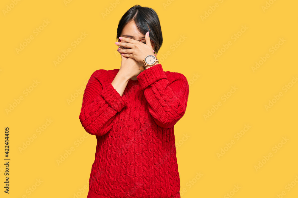 Young beautiful hispanic woman with short hair wearing casual sweater and glasses covering eyes and mouth with hands, surprised and shocked. hiding emotion
