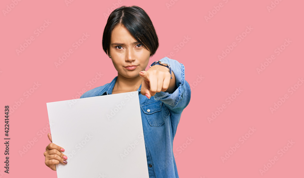 Beautiful young woman with short hair holding blank empty banner pointing with finger to the camera and to you, confident gesture looking serious Beautiful young woman with short hair holding blank empty banner pointing with finger to the camera and to you, confident gesture looking serious