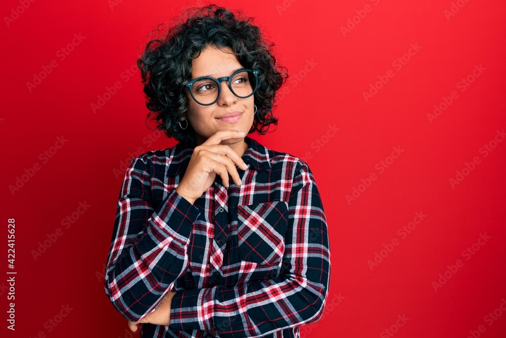 Young hispanic woman with curly hair wearing casual clothes and glasses with hand on chin thinking about question, pensive expression. smiling with thoughtful face. doubt concept. Young hispanic woman with curly hair wearing casual clothes and glasses with hand on chin thinking about question, pensive expression. smiling with thoughtful face. doubt concept.