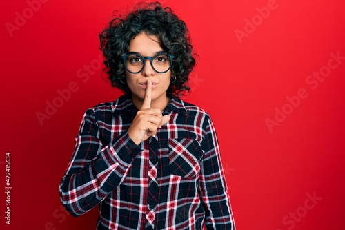 Young hispanic woman with curly hair wearing casual clothes and glasses asking to be quiet with finger on lips. silence and secret concept.