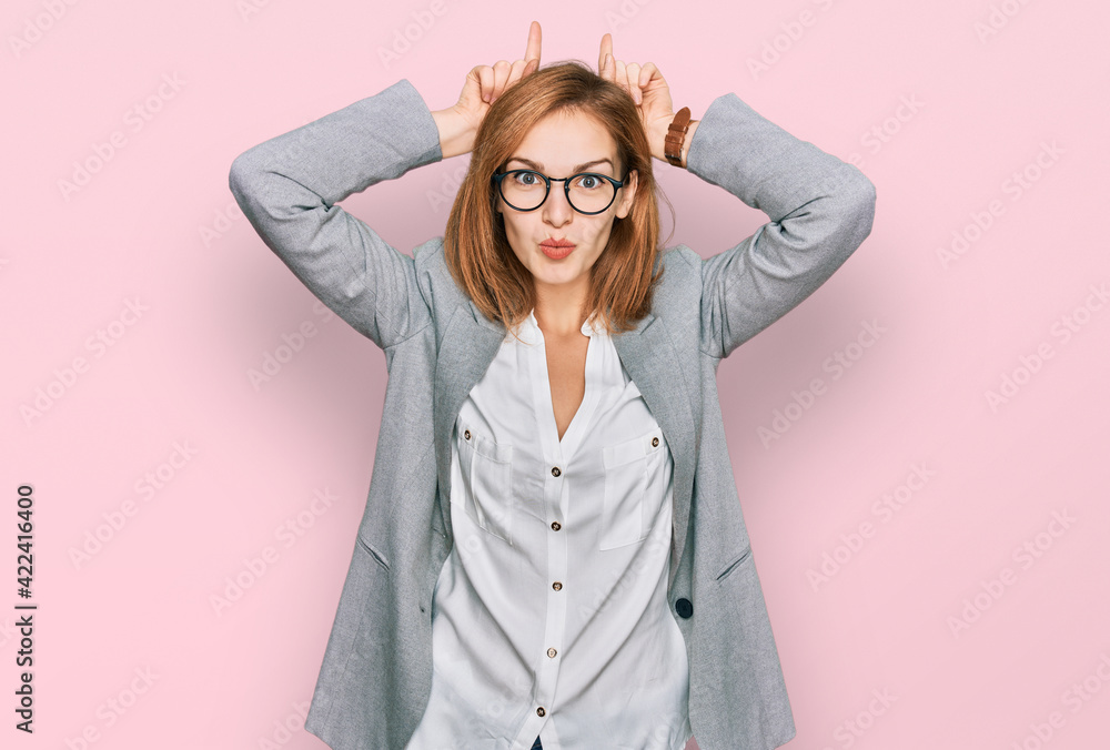 Young caucasian woman wearing business style and glasses doing funny gesture with finger over head as bull horns Young caucasian woman wearing business style and glasses doing funny gesture with finger over head as bull horns