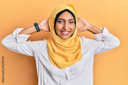 Young brunette arab woman wearing traditional islamic hijab scarf relaxing and stretching, arms and hands behind head and neck smiling happy © Krakenimages.com