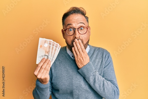 Handsome middle age man holding 10 united kingdom pounds banknotes covering mouth with hand, shocked and afraid for mistake. surprised expression photo