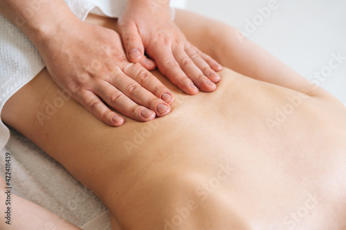 Close-up top view of male masseur massaging lower back of young woman lying on massage table at light spa salon. Experienced chiropractor performs wellness treatments for lady with back pain.