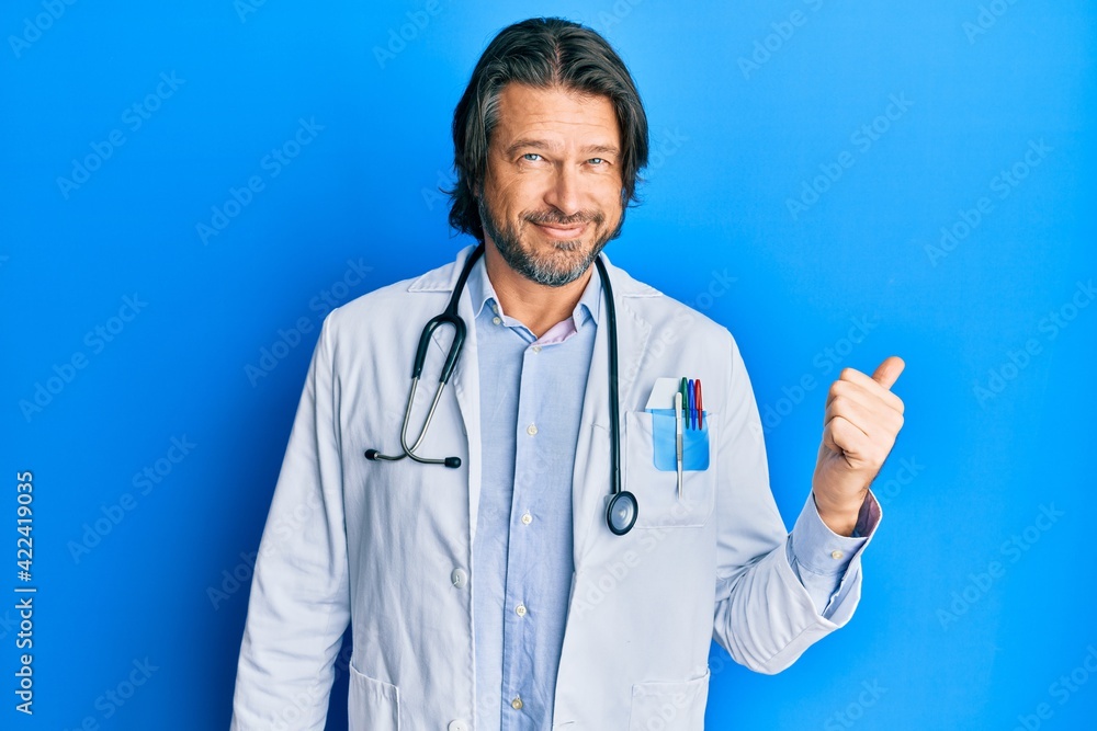 Middle age handsome man wearing doctor uniform and stethoscope smiling with happy face looking and pointing to the side with thumb up. Middle age handsome man wearing doctor uniform and stethoscope smiling with happy face looking and pointing to the side with thumb up.