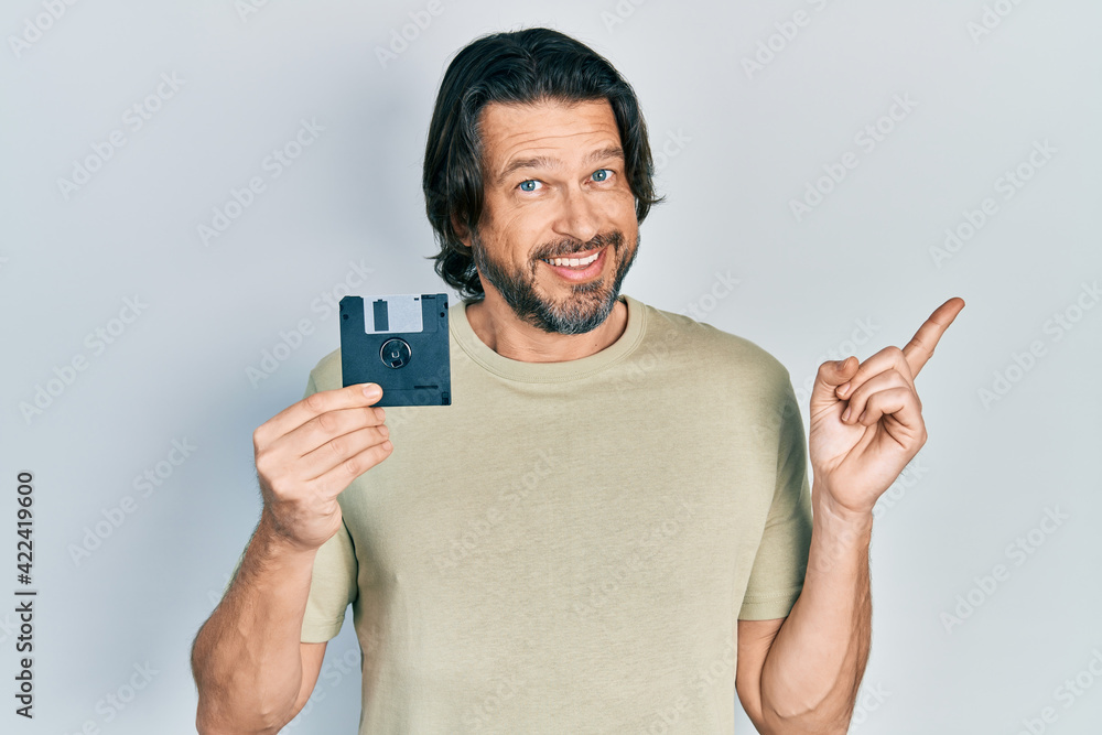 Middle age caucasian man holding floppy disk smiling happy pointing with hand and finger to the side Middle age caucasian man holding floppy disk smiling happy pointing with hand and finger to the side